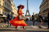 lady with orange dress and black and orange large floppy hat dress is swaying in street black heels she is black and long hair large luxury purse walkingin the streets of paris with the ifle tower in the back ground ppl in the backgound dressed nicely